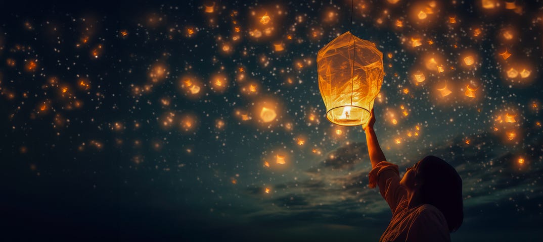 A dark evening with a woman in the foreground letting go of her paper lantern to rise with thousands of other glowing paper lanterns being released into the night.