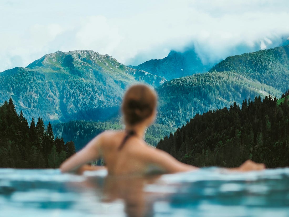 View of the back of a young woman from the shoulders upward standing in a lake with pine trees and mountains in the distant background.