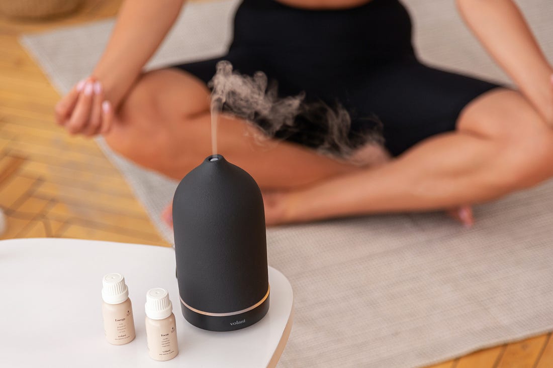 A diffuser on a white table rests in front of a woman pictured from the waist down sitting in a yoga pose on the floor.