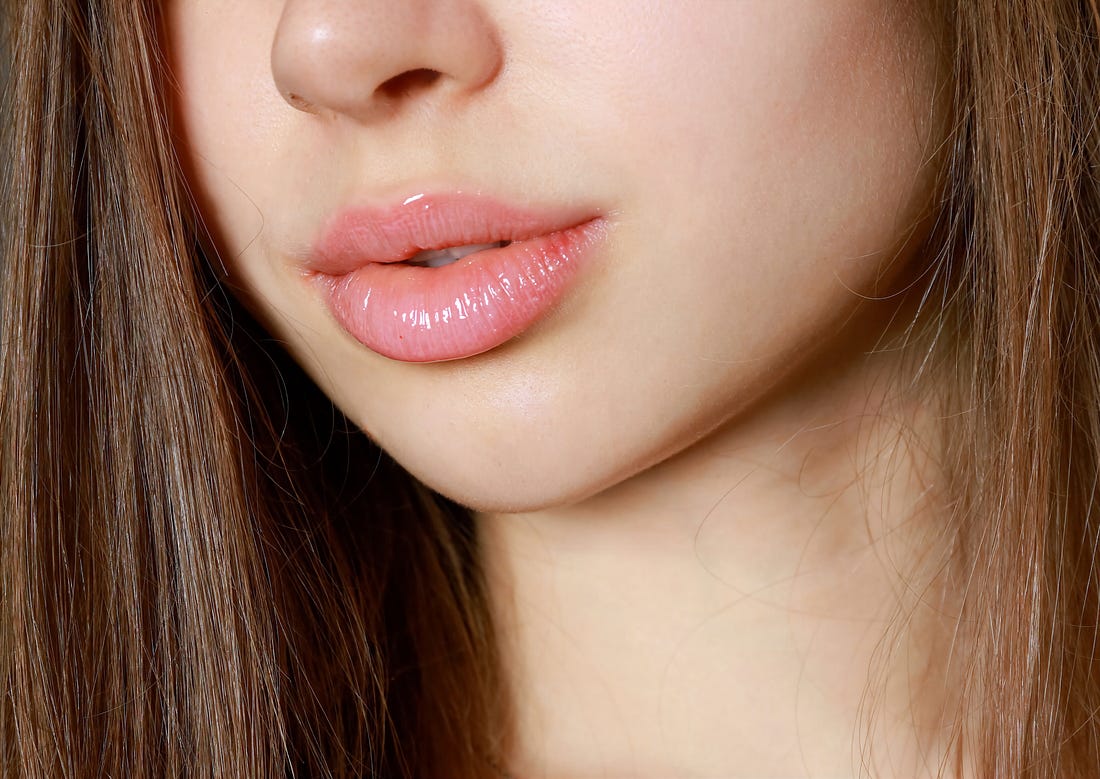 Close up view of a young woman’s face from the tip of the nose to mid-neck area with long hair and full clear glossed lips.