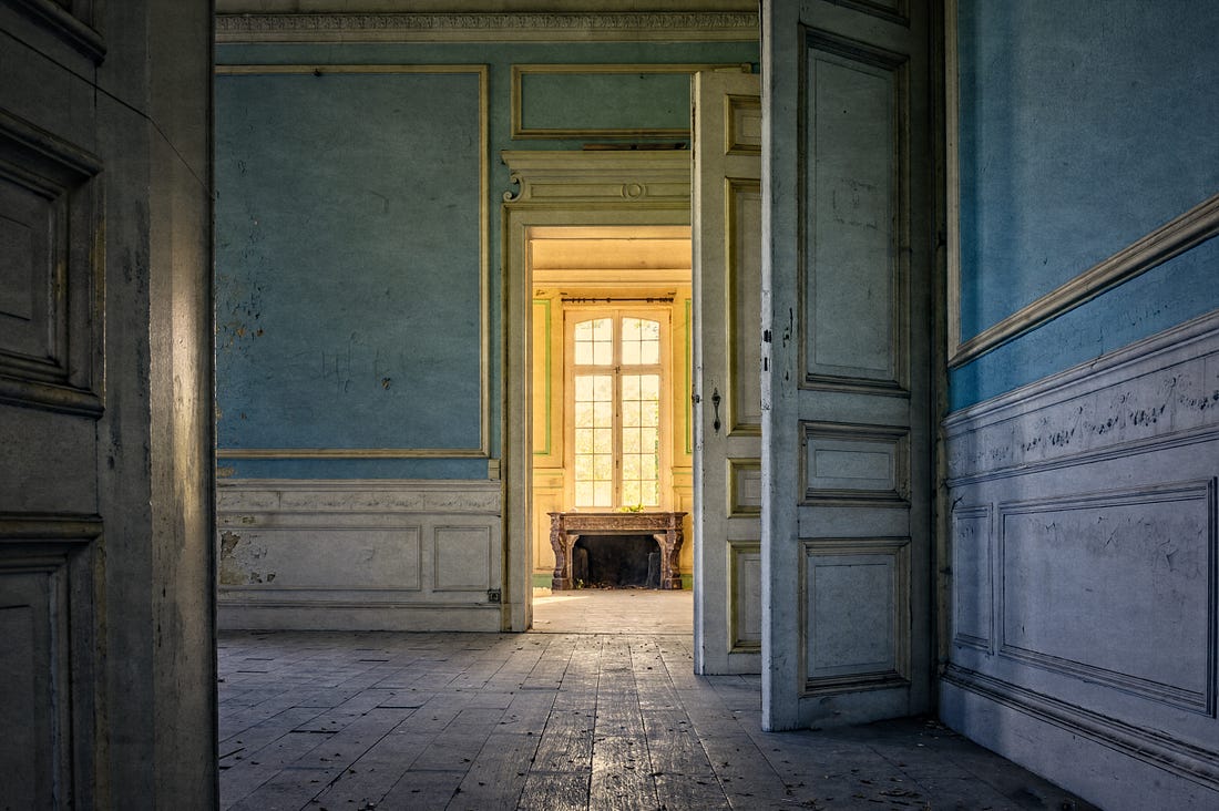Inside view of the large room of an old empty house with peeling paint and wallpaper, and warped hardwood floors.