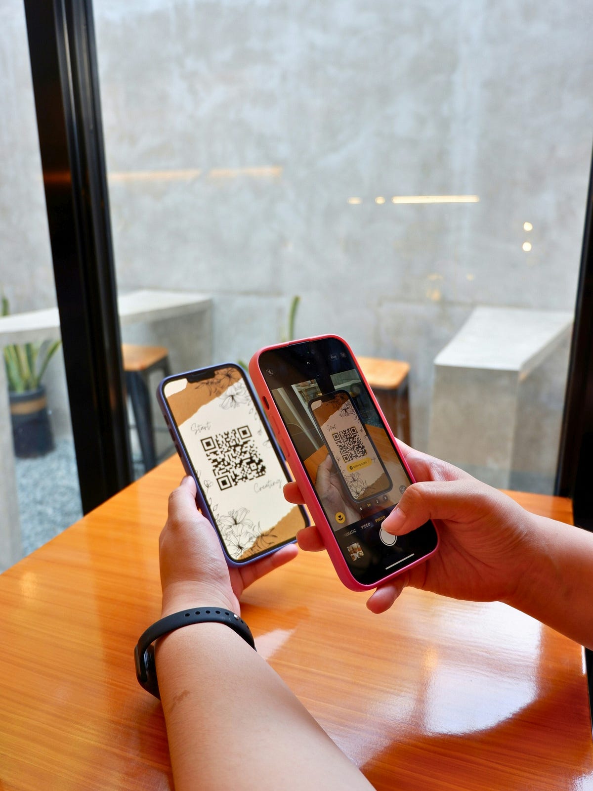 Hands holding two smartphones on a wooden table, with one phone displaying a QR code and the other phone using its camera to scan the code indoors near a window.