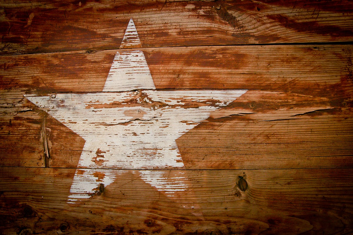 A lone faded creamy yellow-white five-pointed star painted on a piece of multi-toned dark pumpkin-colored grainy weathered wood reminds you of something you would see on a Texas barn.