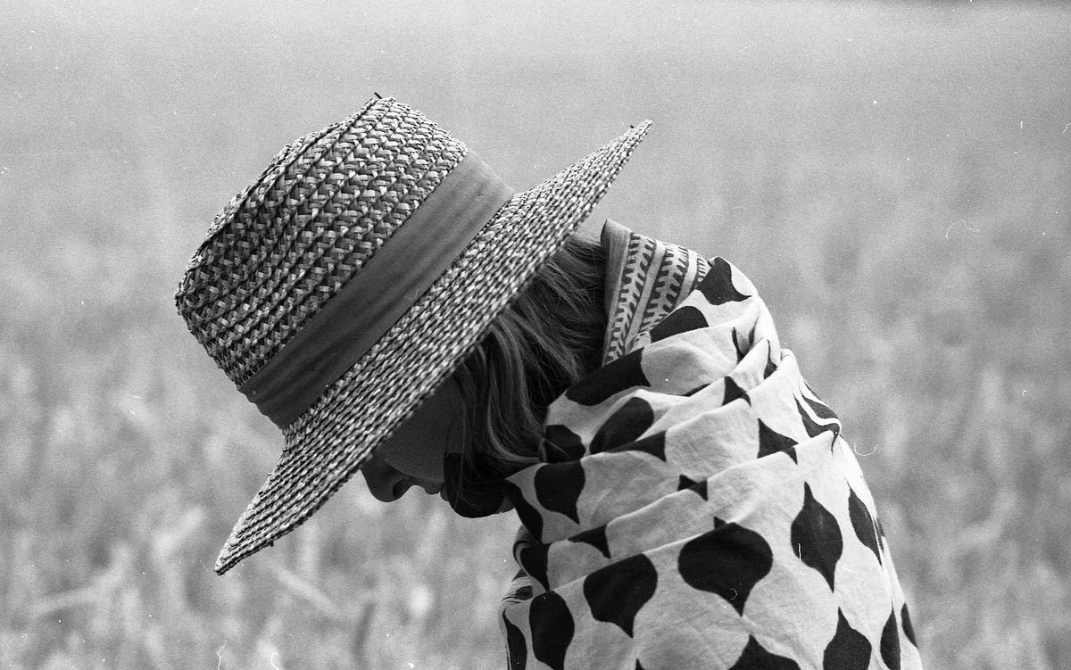 A side view of a man with his head down as if humbled with shame stands in front of a bright white background. He is wearing a white shirt detailed with black teardrop-like random shapes and a wide-brim woven straw hat with a dark gray border about two inches in height at the base of the crown above the brim.