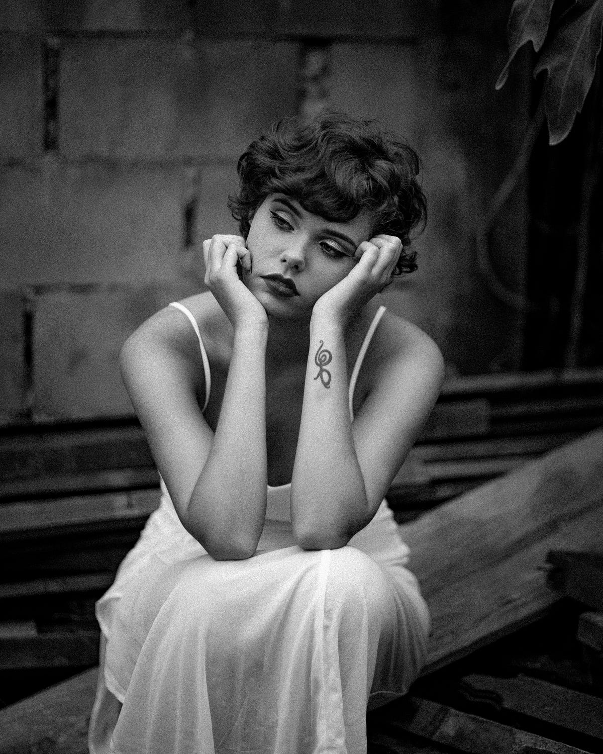 grayscale photo of a beautiful woman sitting on wooden planks while resting her head on her hands