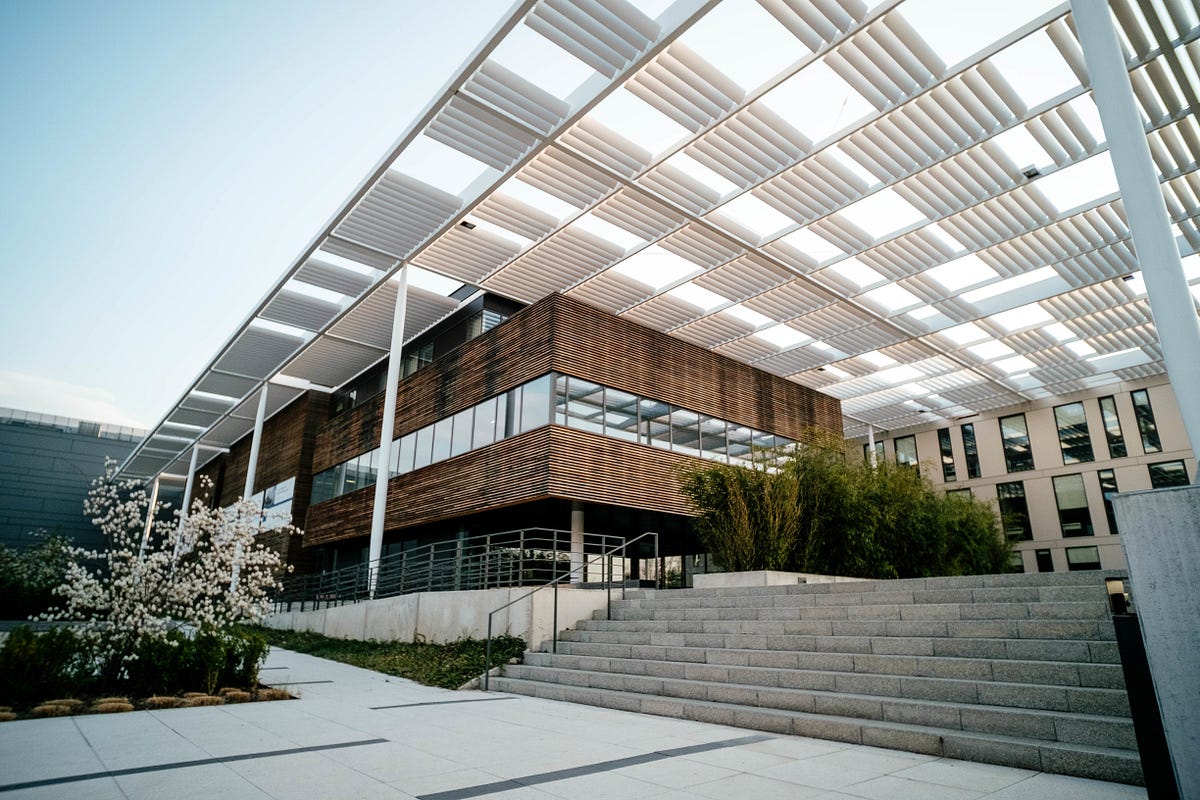 A striking example of contemporary architecture, this building features clean lines, natural wooden cladding, and expansive glass windows. A geometric white canopy adds visual rhythm while filtering sunlight, enhancing energy efficiency. Surrounded by greenery and open spaces, it blends modern design with functionality, creating an inviting and sustainable environment for work or learning.
