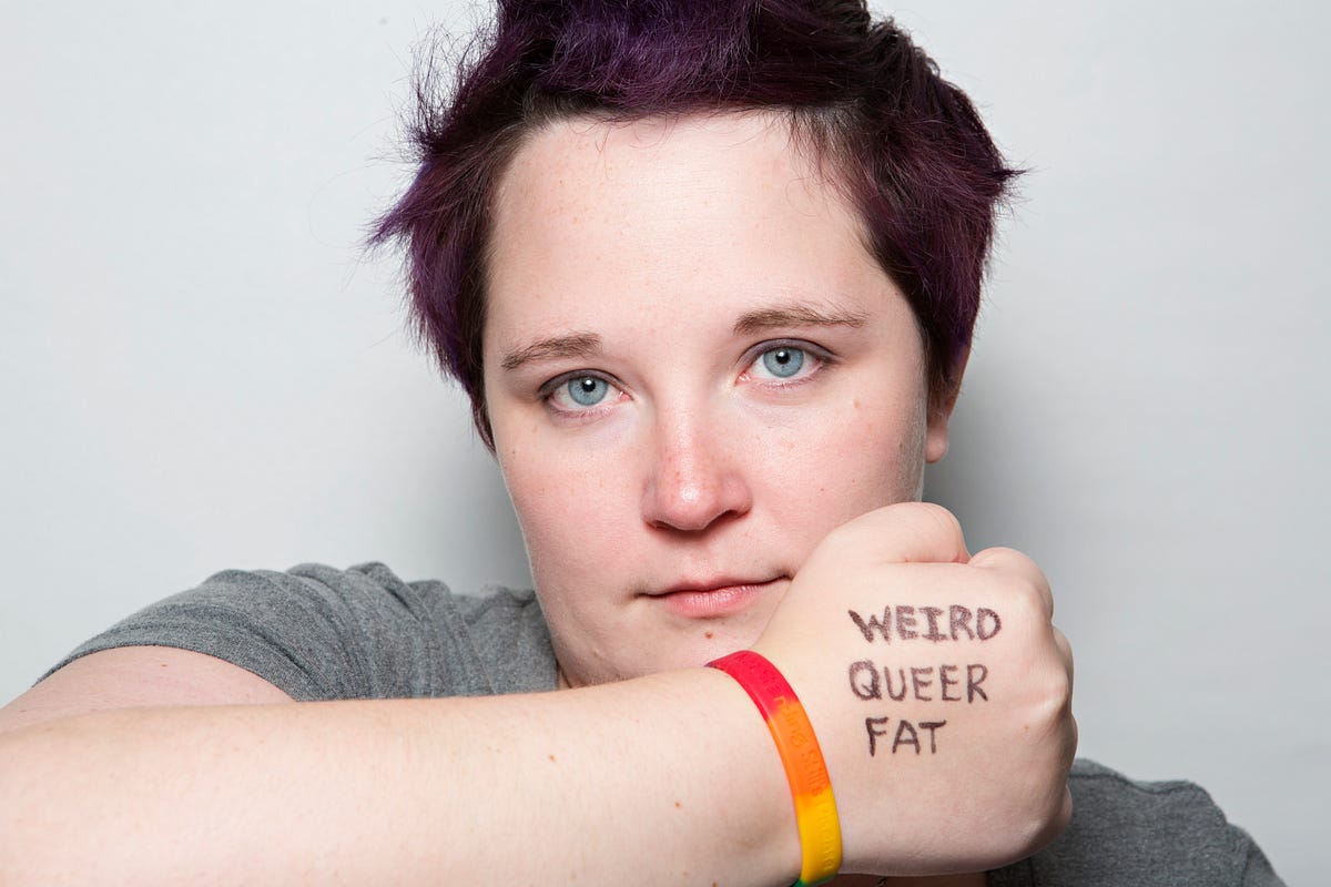 [2016: a photo of Kirsten holding her right arm up in front of her mouth with her hand in a fist, back of the hand facing out with the words “Weird Queer Fat” written on it in black sharpie; a rainbow bracelet is on her wrist; she is making a neutral, though kind of sad, face with her blue eyes staring out piercingly; she has on a grey tee and has dark brownish purpleish hair spiked into a faux hawk]