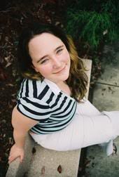 [2005, high school senior picture: Kirsten sits on a stone bench with white pants and a teal/dark blue striped collared shirt; she is looking up towards the camera while smiling; she has long hair that goes from dark brown to blondeish at the ends]