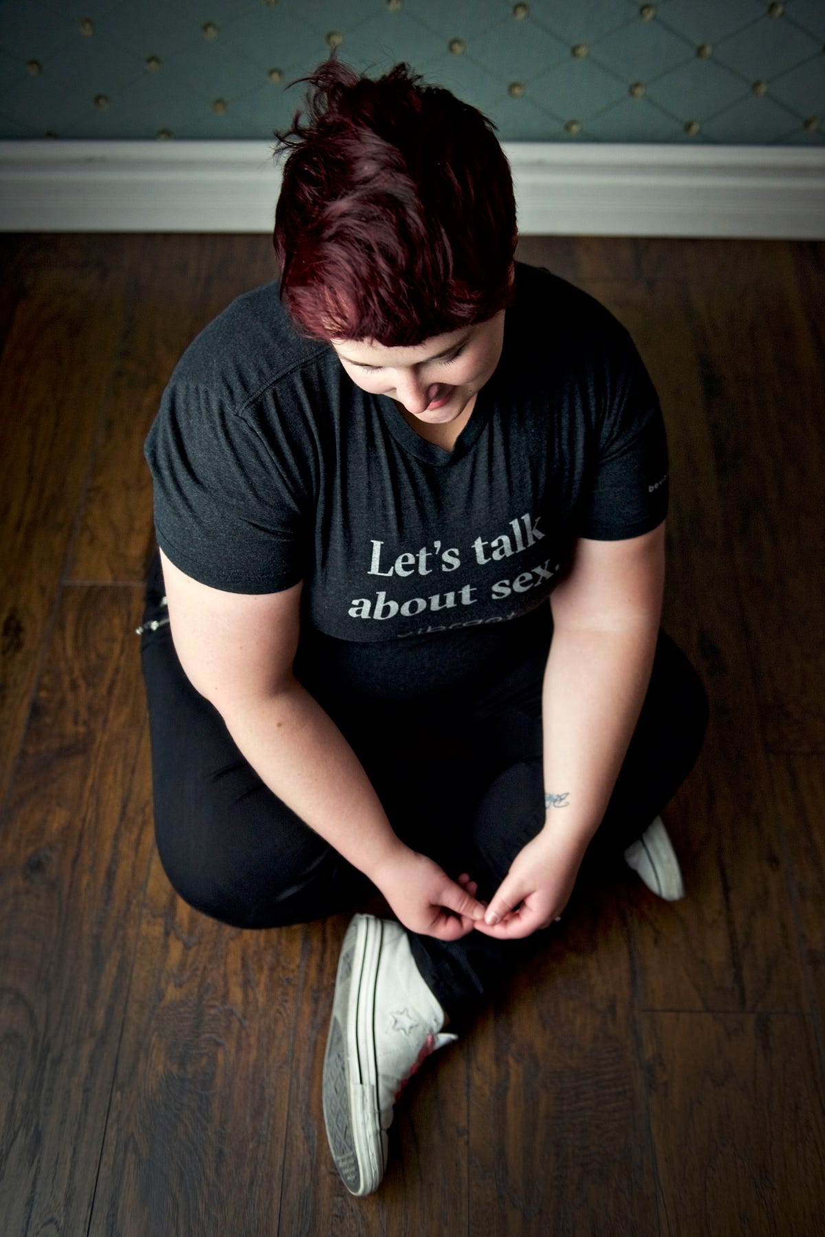 [2017: pic of Kirsten from above, sitting on a dark wooden floor with white moulding and a blueish background; she has her legs bent criss-cross style, though not completely, and is looking down at/playing with her hands; she has on black jeans, a gray tee shirt with white text “Let’s talk about sex — Vibrant” and her white Converse are visible; her hair is reddish-purpleish]