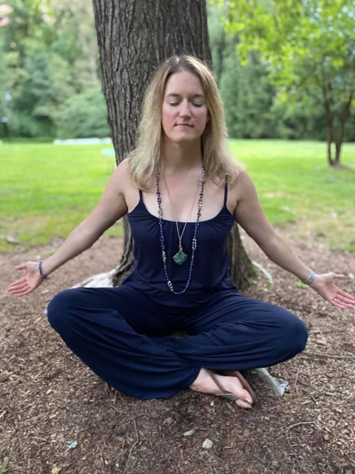 Woman with flowing long blonde hair sitting cross legged in front of a tree with arms extended behind her