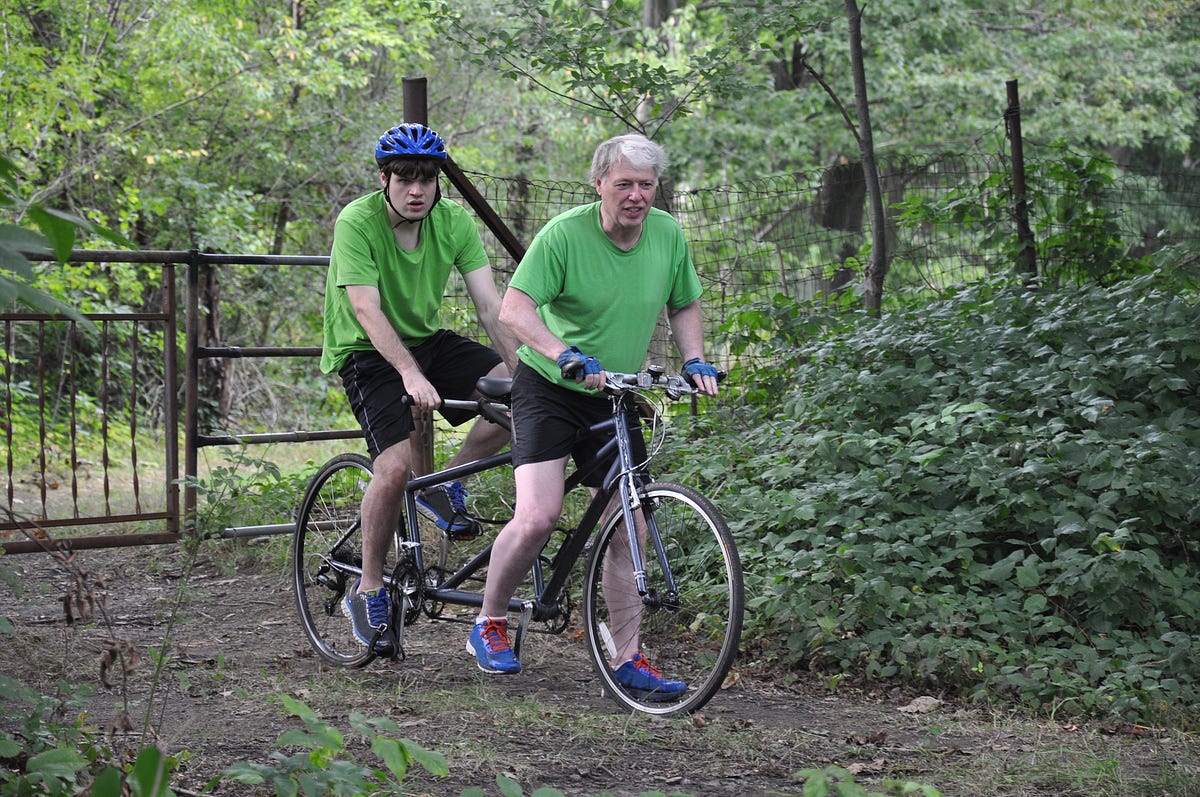Homem branco de cabelo grisalho com um jovem branco autista de capacete azul, ambos em cima de uma bicicleta em um parque.