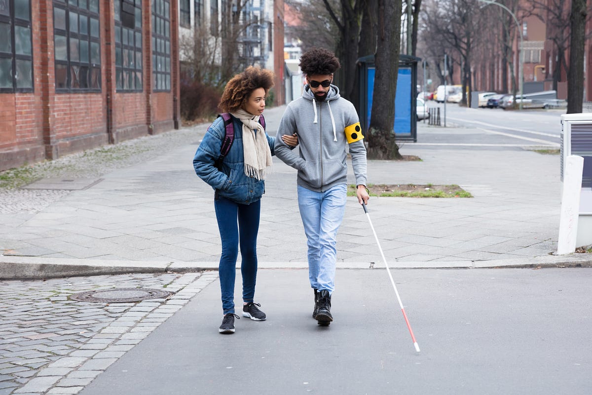 Mulher negra acompanhando um homem negro e cego, que usa óculos escuro e bengala, andando pela rua de uma cidade grande.