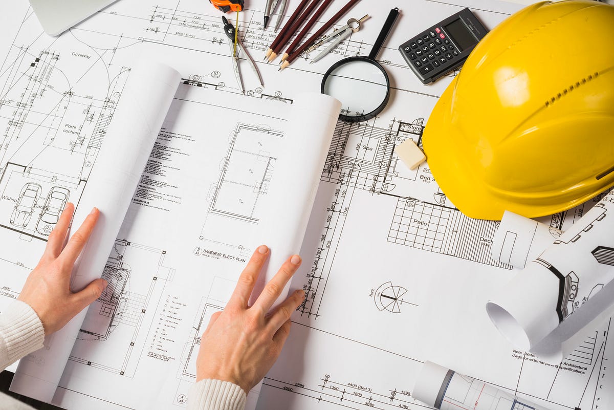 A detailed architectural workspace featuring blueprints, design drawings, and essential tools for planning. The setup includes a yellow safety helmet, pencils, calculator, compass, and magnifying glass, symbolizing precision and safety in construction. Hands unfold the floor plan, showcasing technical details for residential and structural design. This image represents architecture, engineering, and the collaborative process of building modern spaces.