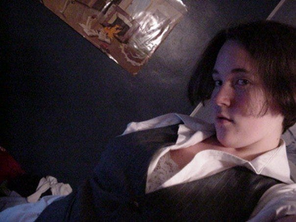 [2006/2007, Freshman in college: Kirsten is taking a selfie; she is wearing a white button up dress shirt with a grey waistcoat and white lacy bra; she has short dark hair and is standing in front of a dark blue wall with an abstract art poster]