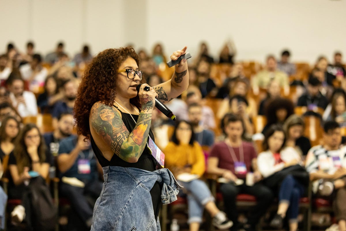 Mulher de cabelo crespo e tatuada falando ao microfone, e ao fundo uma platéia assistindo a palestra.