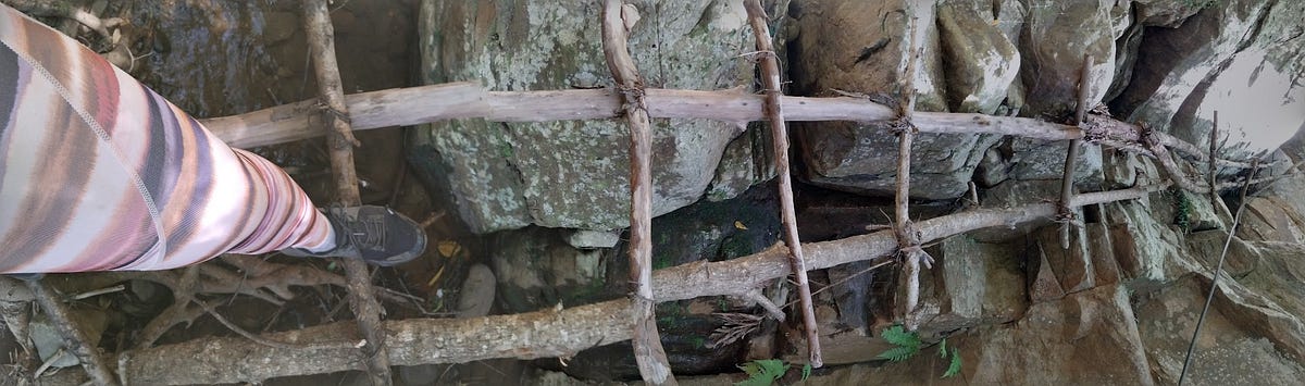 A leg dressed in pink striped yoga pants and wearing black shoes resting on a home-made wooden ladder, set against boulders