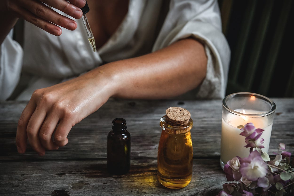 Close up of a person’s arm and hands. They are sitting at a distressed wood table with a lighted candle and two small bottles, holding a dropper with their left hand while applying a drop of oil on their right wrist.