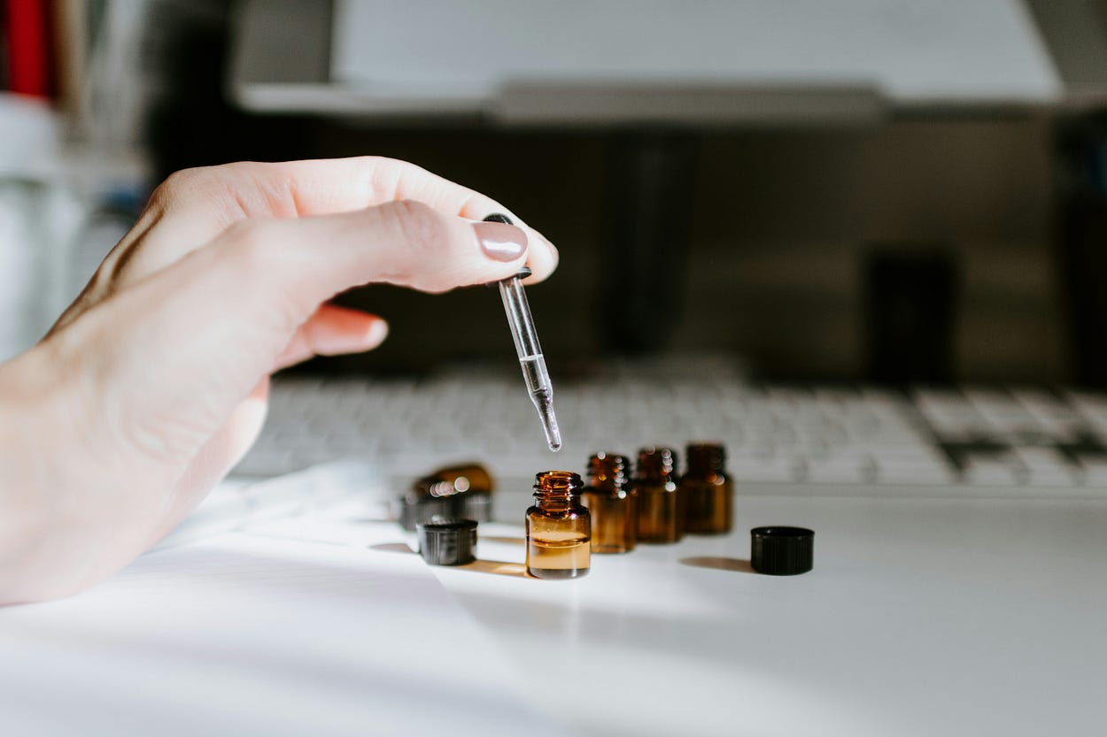 Four uncapped mini-amber bottles on a table. A hand is holding a dropper adding drops to one of the bottles. The background is blurred.