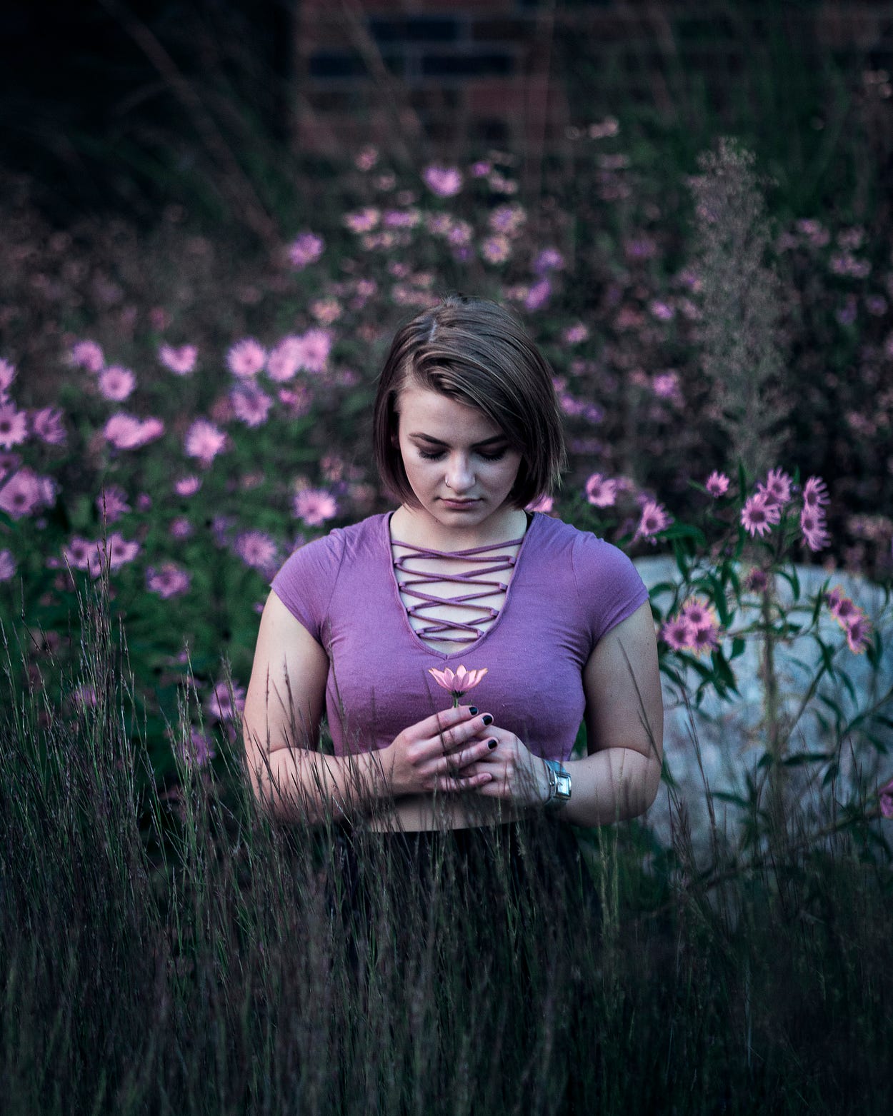 A young woman with a peaceful expression stands in a patch of purple wildflowers looking down at a single flower stem held upright in their hands.
