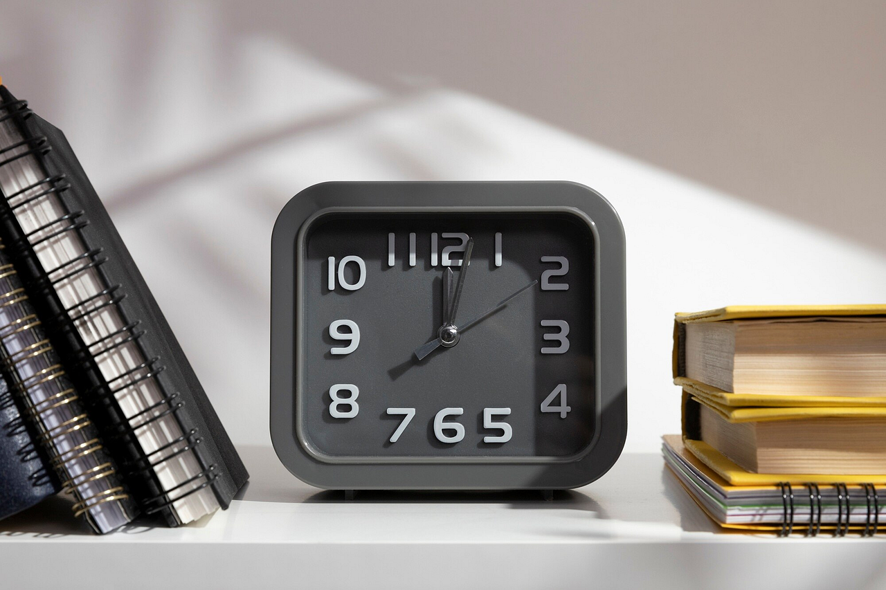 Close-up of a modern gray alarm clock placed on a desk next to books and notebooks, symbolizing productivity and time management. Ideal for a focused study session or a well-planned day.