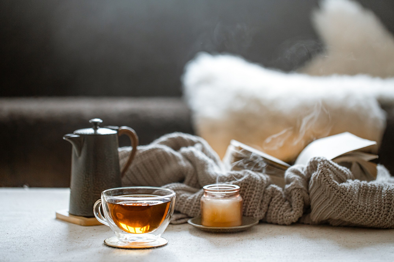 A cozy setup featuring a steaming cup of tea, a flickering candle, and a soft knit blanket — perfect for a calm, reflective morning or an inspiring daily reminder to slow down and savor life’s simple pleasures.