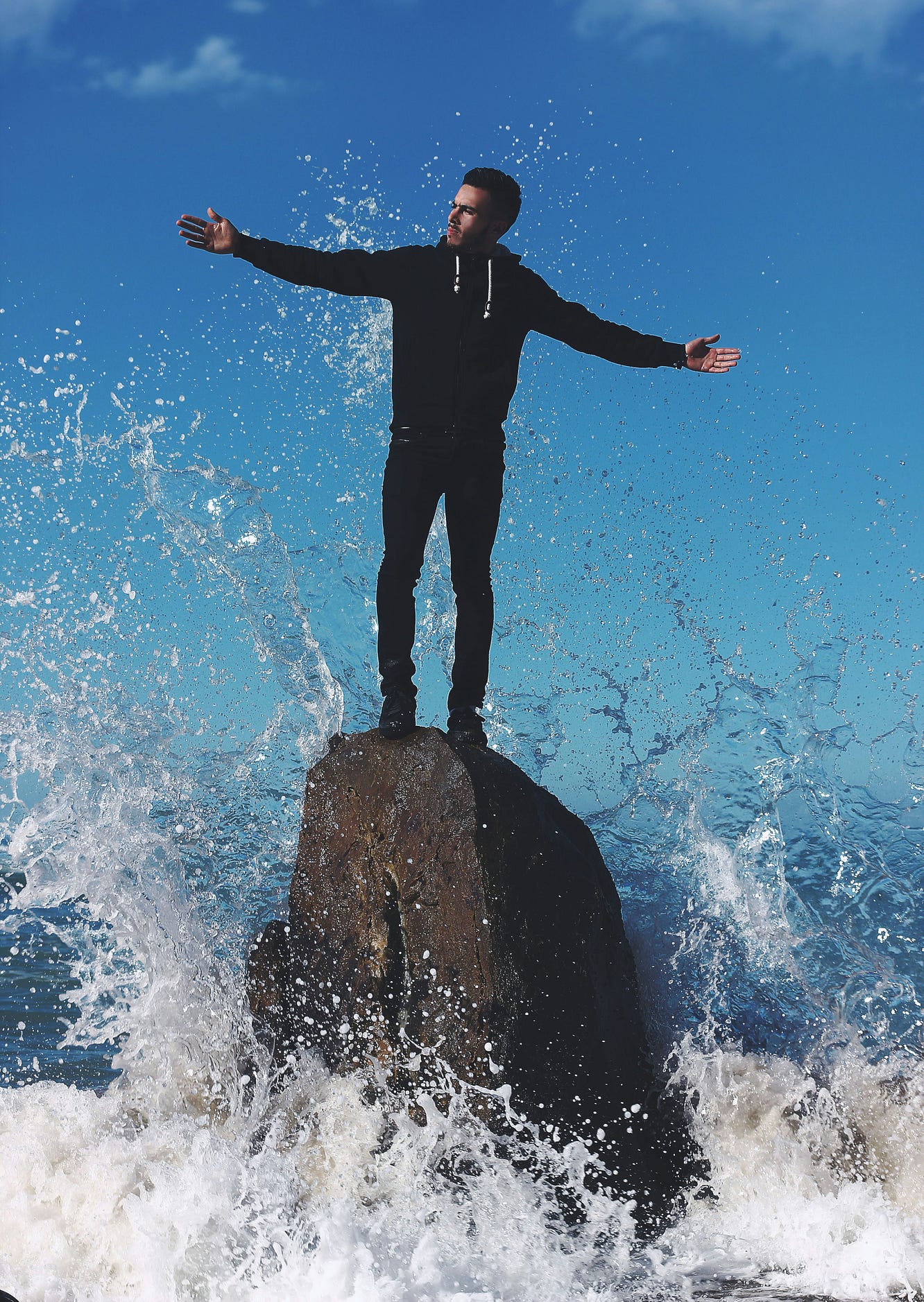 Image of a dressed man standing on a rock with ocean waves beating around the base of the rock.