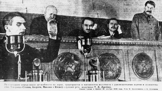 Pseudoscientist Fadim Lysenko speaks at the Moscow Kremlin, gesturing dramatically; behind him in a gallery sit three bored-looking men and, standing, Soviet dictator Joseph Stalin.