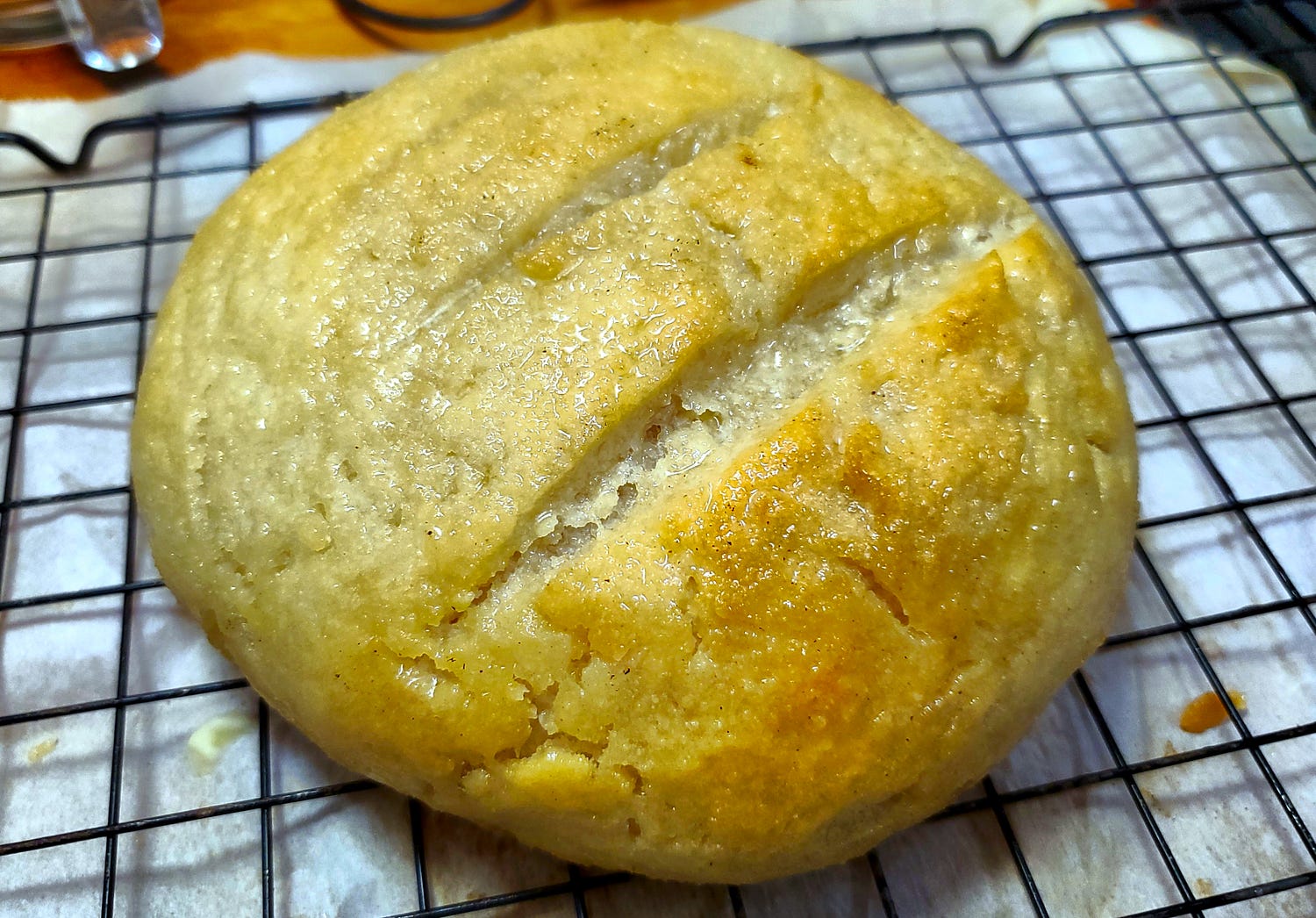 A round loaf of bread with three slits on the top is sitting on a baking rack cooking over a piece of parchment paper.