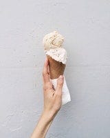 A hand gripping an ice cream cone with 2 scoops of frozen dessert against a white backdrop.