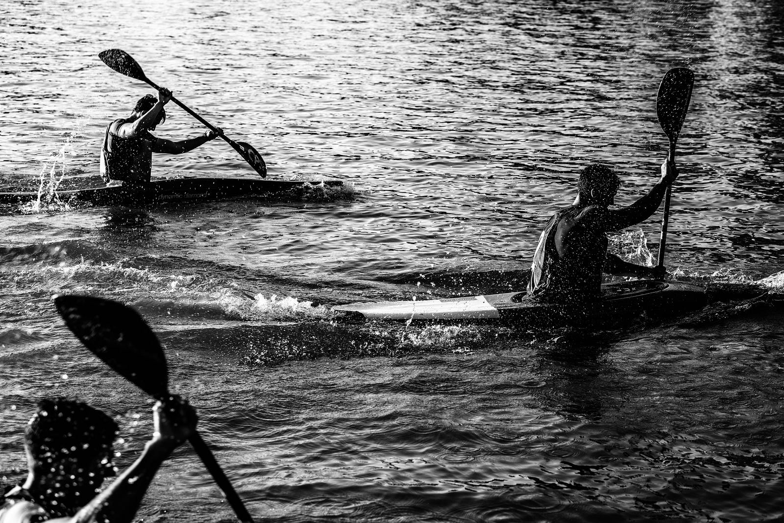 Black and white image of three kayakers working hard on flat water. They are owning the suck.