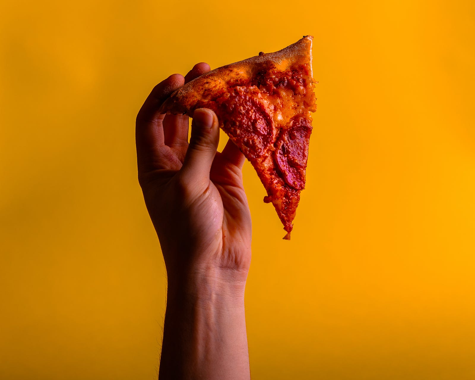 A hand extends from the lower image to hold a single piece of pizza. The slice dangles downward. The background is a mustard color. To reduce weight, exercise alone is not adequate to drop weight; dropping calories is a must.