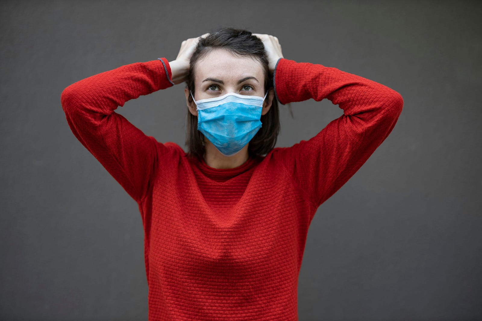 A young woman places her hands on the top of her head as she dons a blue paper surgical mask.