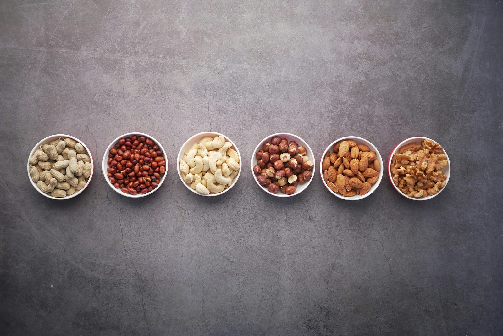 Six small white bowls of different types of nuts are arrayed left to right.