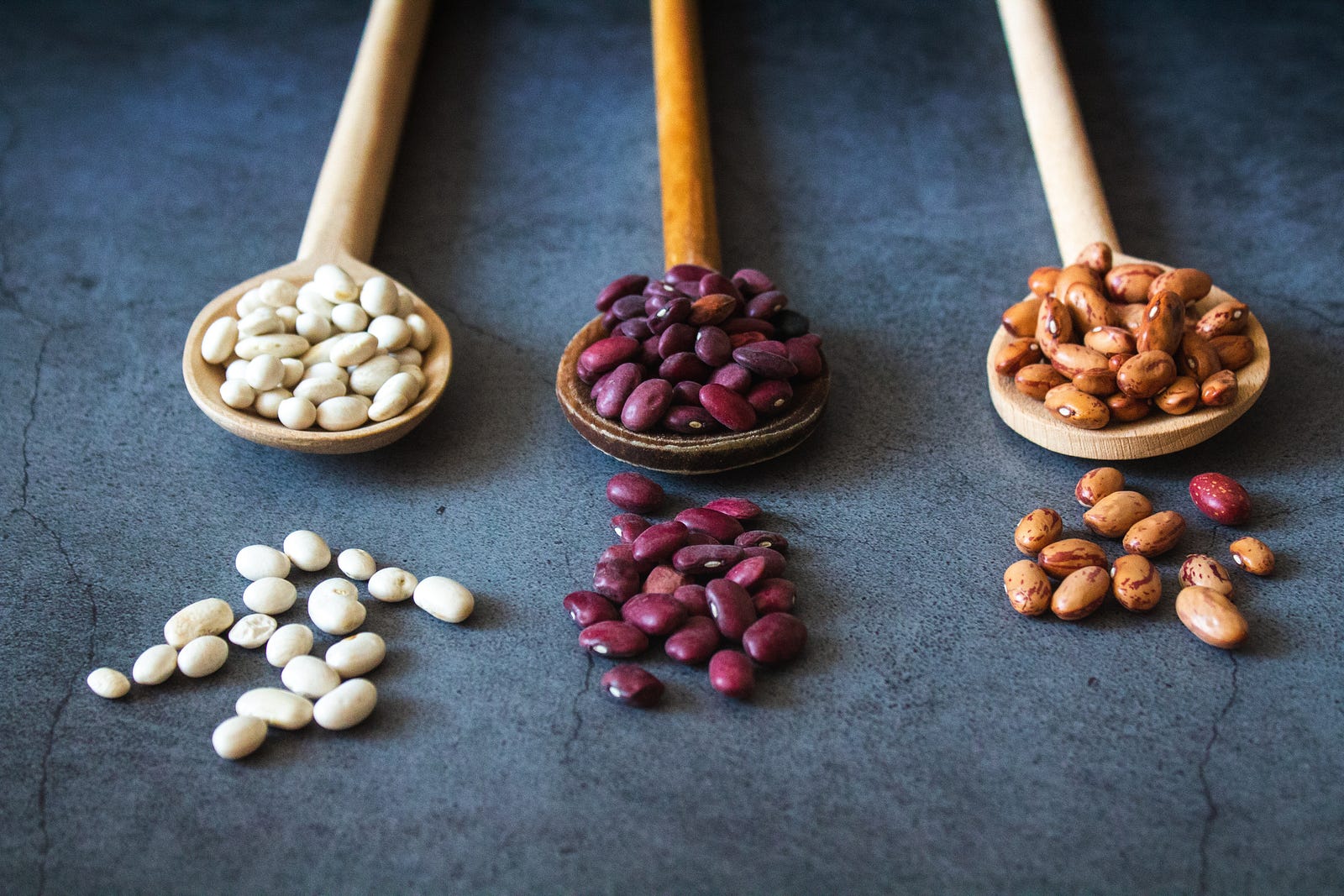 Three small wooden spoons extend from the image top, holding white, purple, and brown soybeans, respectively. The table is blue-gray. Soy is rich in plant estrogens, known as isoflavones.