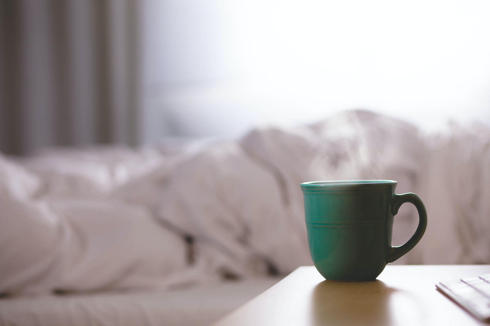 A green cup (with steam rising out of it) sits on a desk in the foreground. There are messy sheets on a bed in the background. U.S. hospitalization rates from RSV and flu are rising, and outpatient visits for respiratory illnesses are abnormally high, according to CDC data.