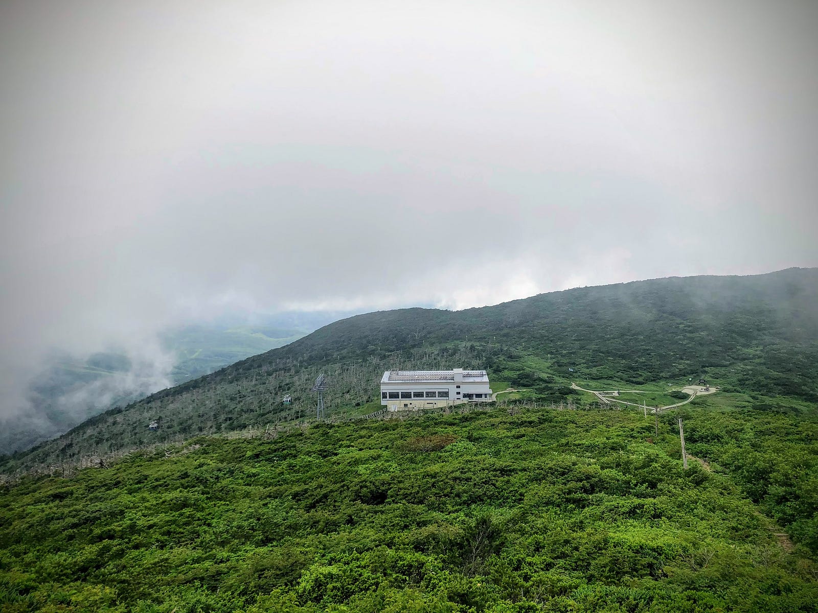 The white Zao Ropeway Jizo Sancho Station amongst a sea of green and a white sky, seen from the summit of Jizo-dake, with statues of Jizo-san to the right.