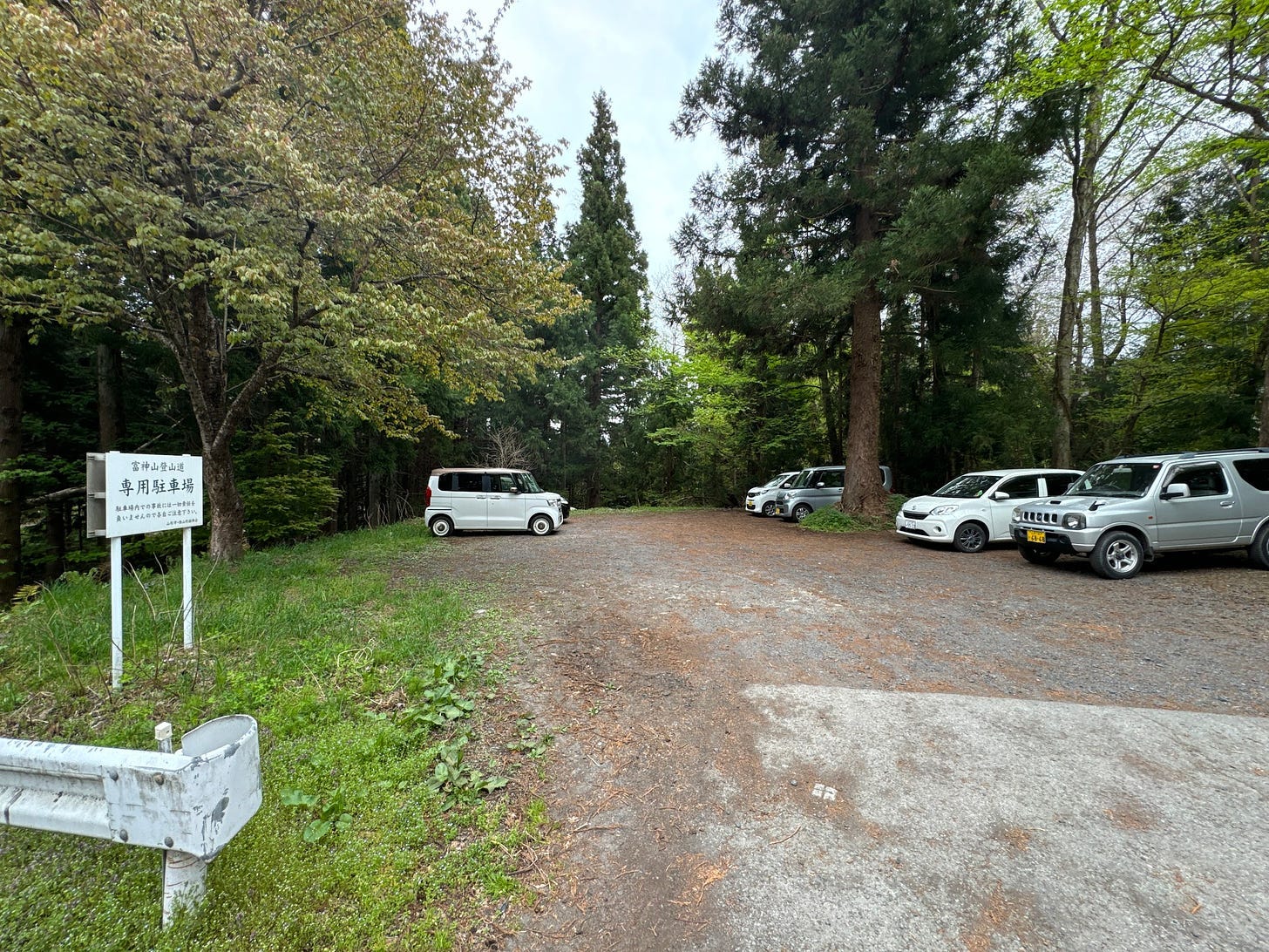 A car park at the Shindo Trailhead to Togami-yama. Cars parked in a gravel clearing under towering trees of all shades of green.