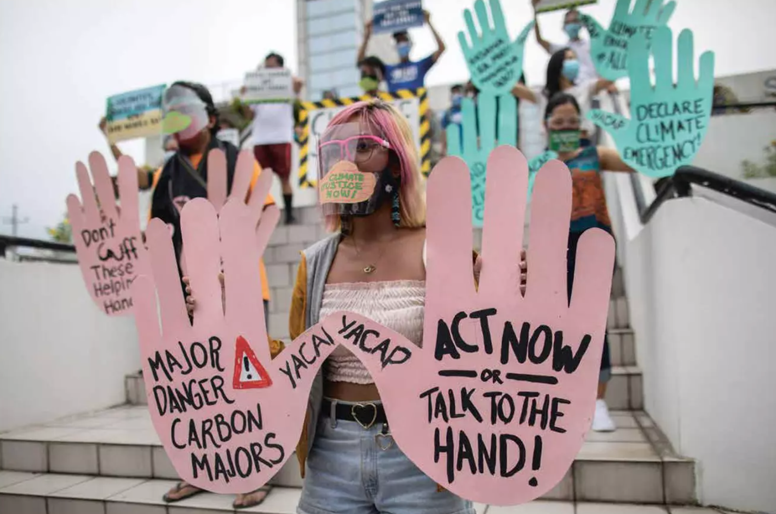 The image shows a woman stood in a croud of protestors wearing a face mask and glasses, holding up two big pink hands which say 'Major danger Carbon Majors' and 'Act Now or Talk to The Hand'.