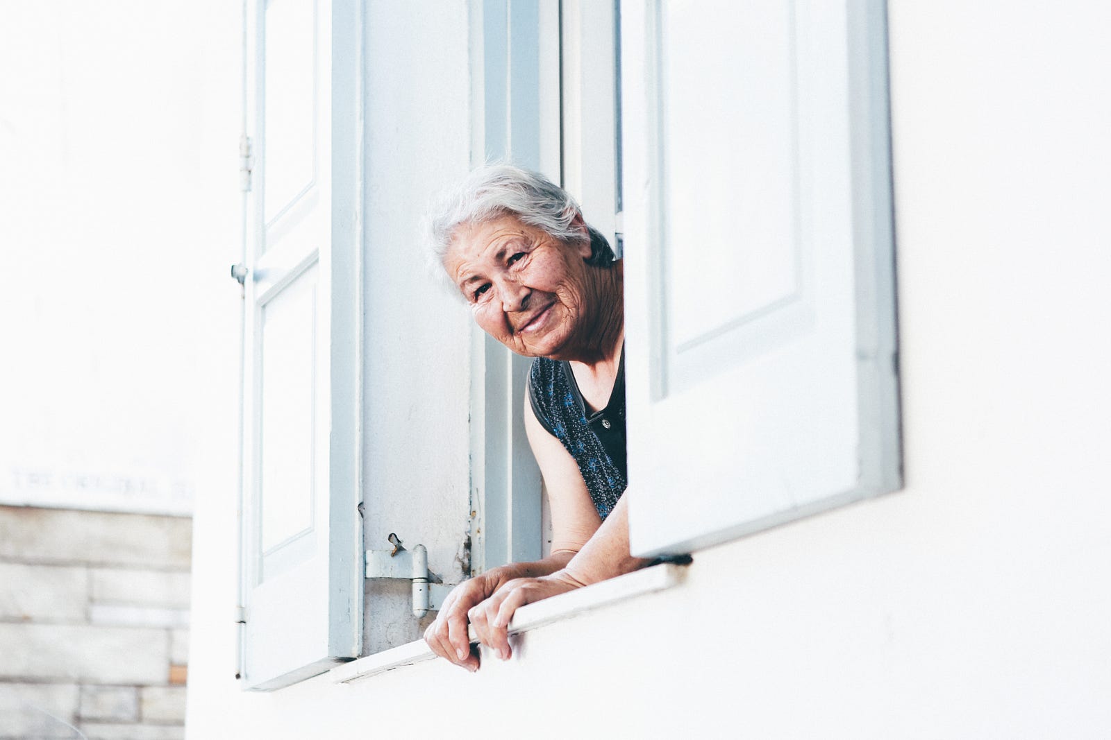 An older woman leans out of a window of a white house. She is percehed between two white shutters.