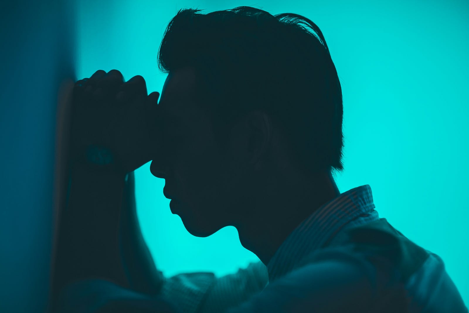 A man looks in pain as he leans forward against a wall, hands up to his face. Lateral view.