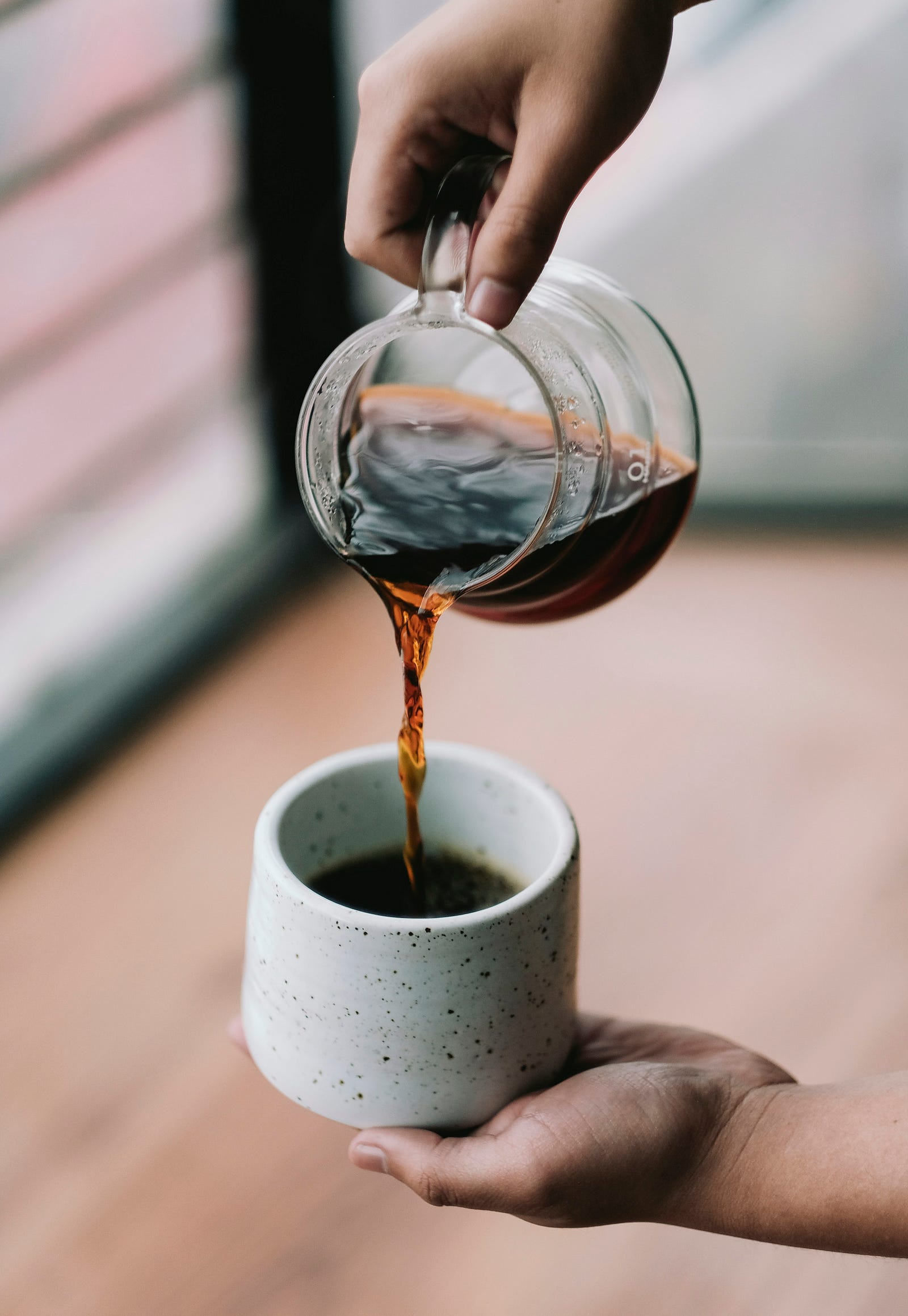 A person’s arm extends to pour coffee from a small glass carafe into a white cup.