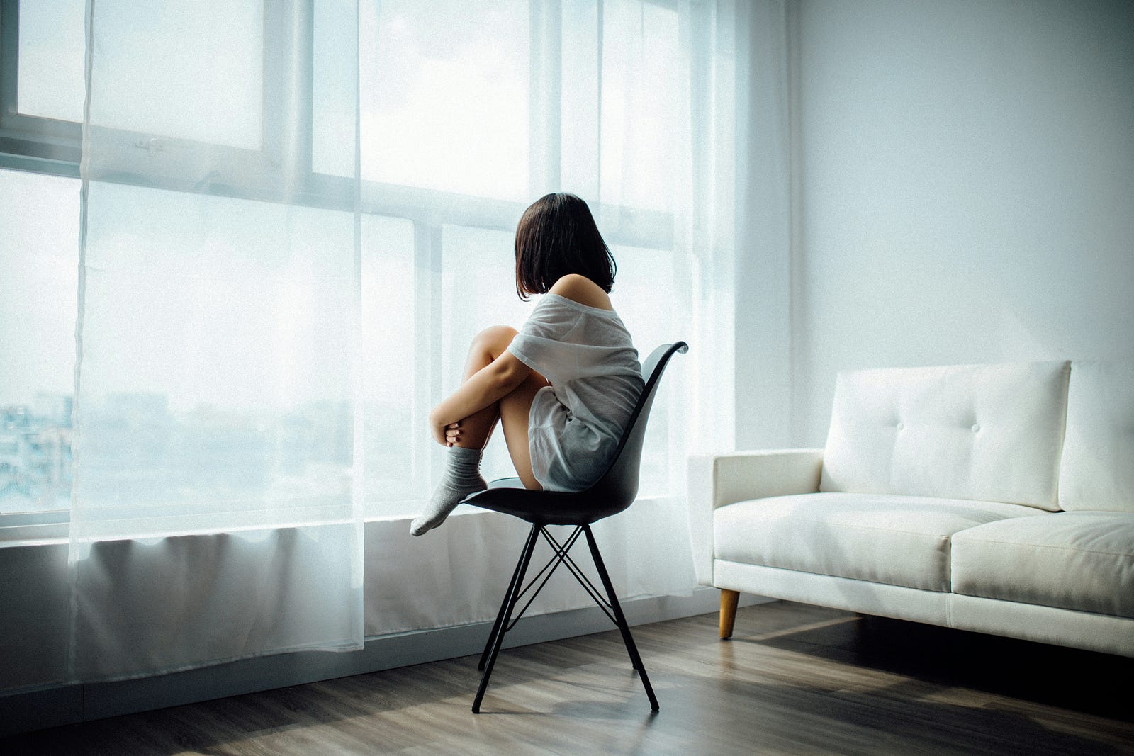 A woman sits in her room, gazing out thought a window.