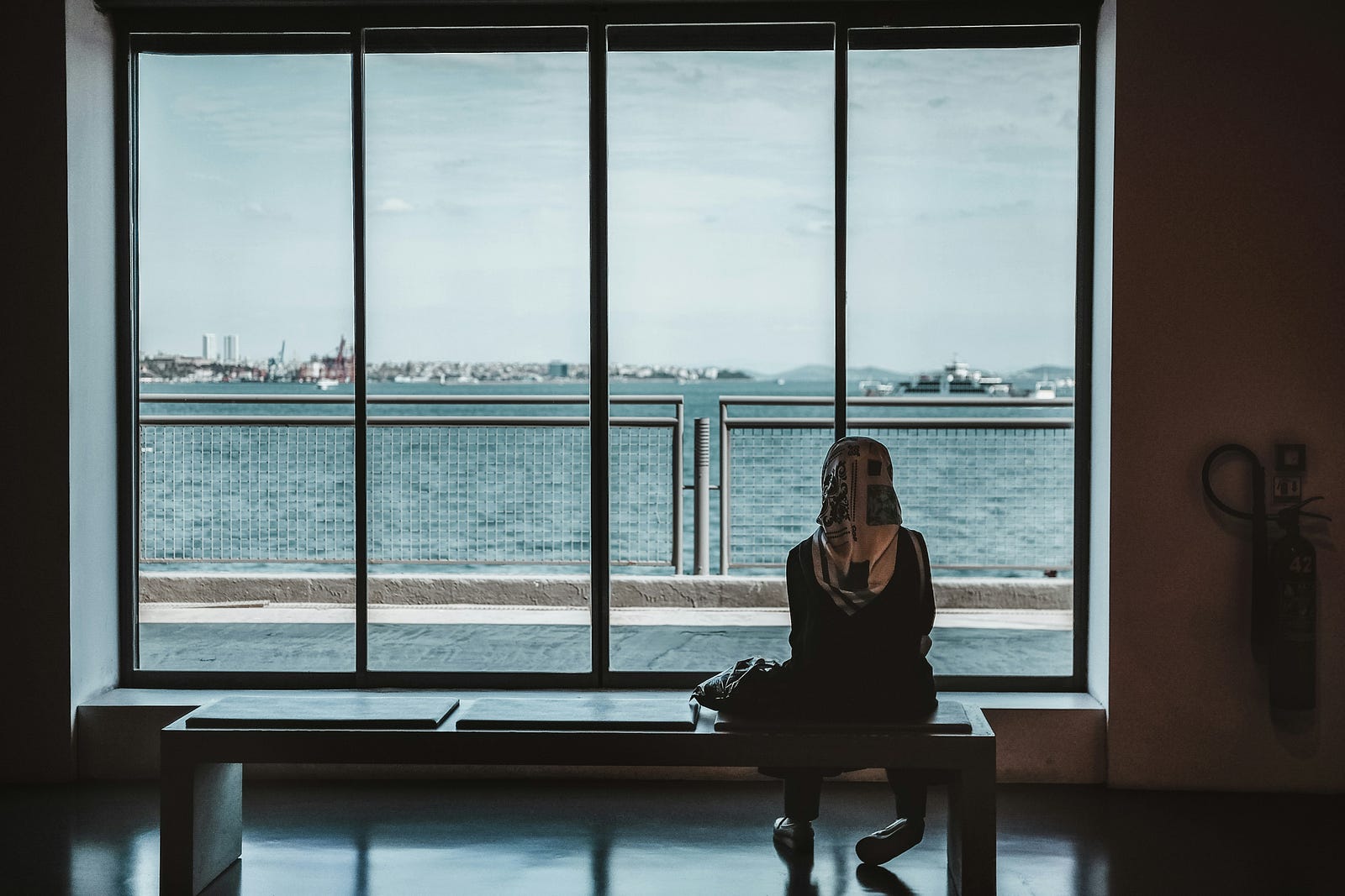 A person sits on the right end of a bench as she peers outside of a large window. We see water in the distance.