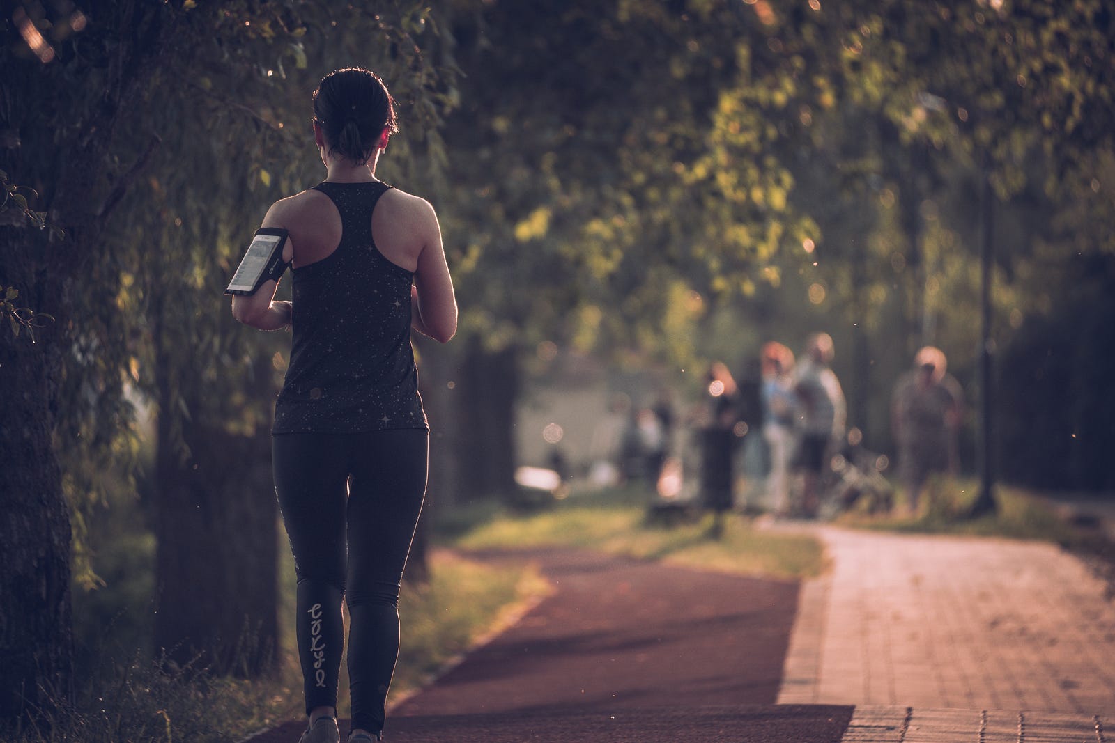 A woman runs away from us on the left side of the image. She jogs on a path with trees on either side. Running outdoors allows an individual to encounter various terrains and can be best for race training.