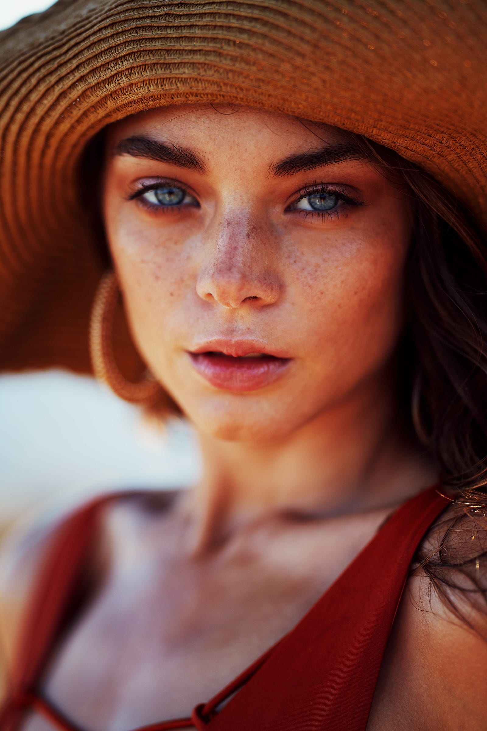 A beautiful, freckled woman looks at us with a neutral expression. She dons a straw hat, has blue eyes, and large wooden hoop earings. Solar exposure is the leading cause of skin cancer, including melanoma.