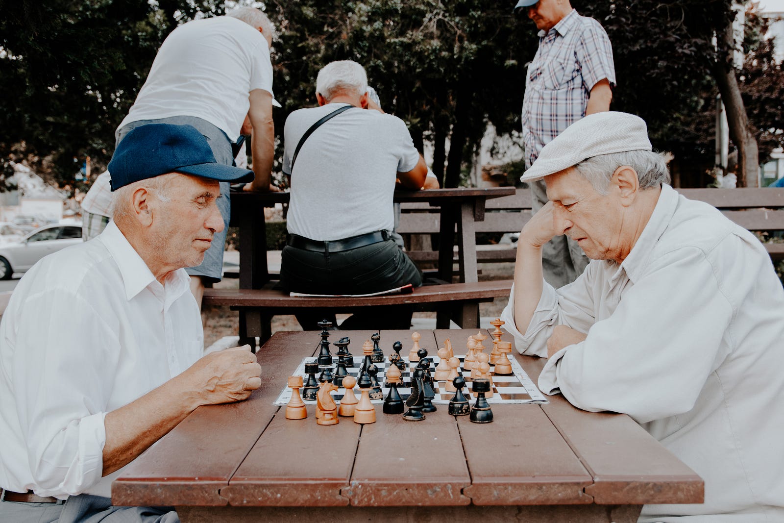 Two older men (when on the left of the screen, and one on the right) play chess in a park. Others play in the background.