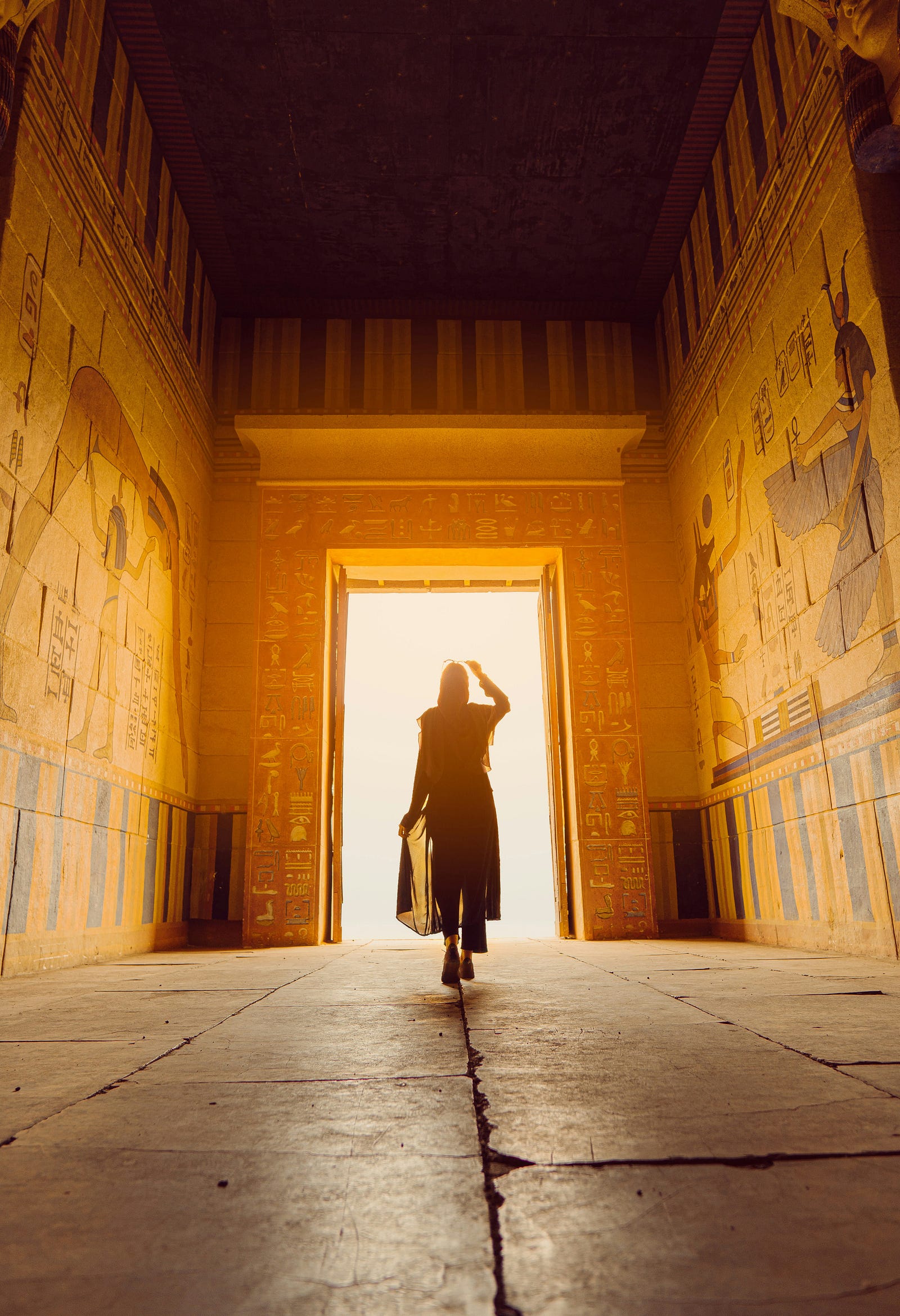 A woman walks away from us, towards the exit door of an ancient Egyptian building.