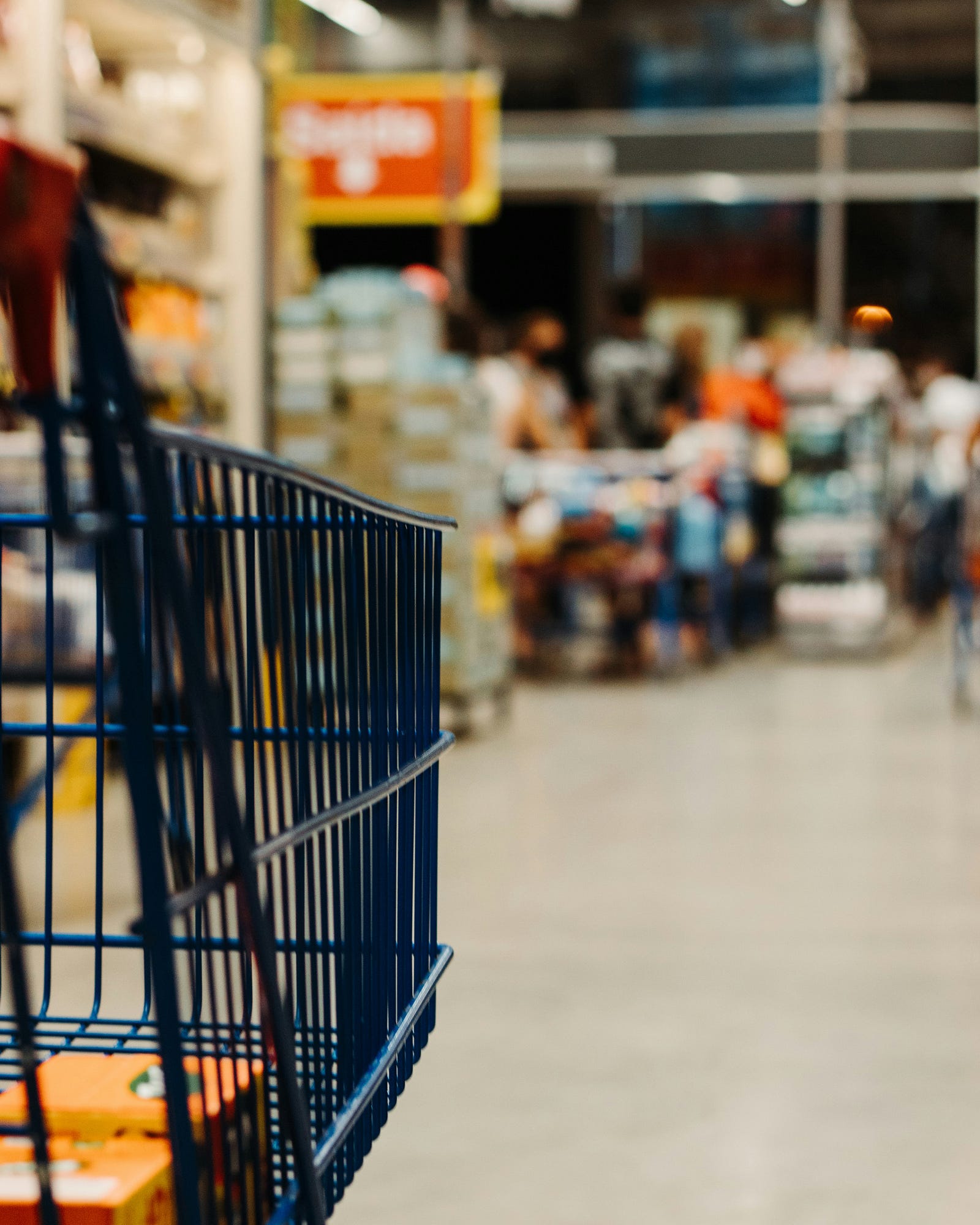 A black shopping cart is in the left foreground. In the background are blurred grocery products. According to a 2012 study published in the International Journal of Food Protection Trends, shopping cart handles are hotspots for various bacteria, including potential nasties like E. coli and Salmonella.