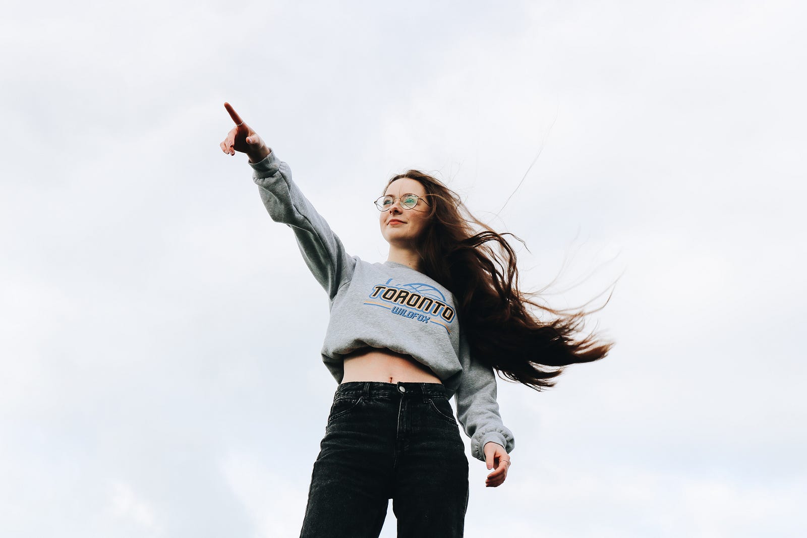 A young woman, clad in a gray sweatshirt and black jeans, points with her right index finger upward at a 45 degree angle. Running can increase confidence.