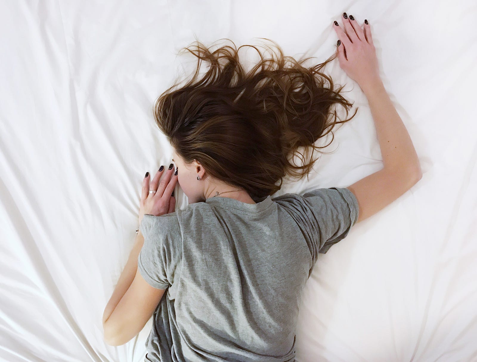 A young brunette lies prone on a bed with a white sheet. Her left arm is folded back against her face. Her left arm extends up. A new study shows that individuals taking daytime naps had significantly improved cognitive performance compared to those who stayed awake.